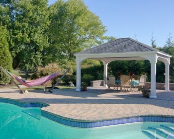 Traditional white vinyl pavilion with gray asphalt shingles on a brick patio by the pool.