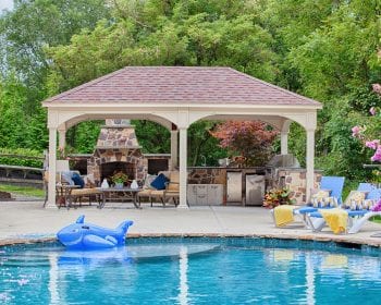 Patio furniture and outdoor kitchen underneath a traditional ivory vinyl pavilion by the pool.