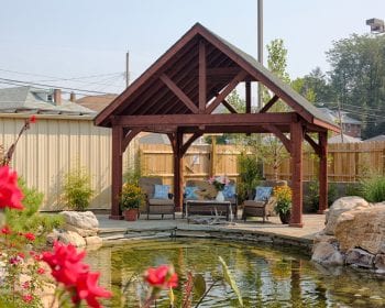 Alpine cedar wood pavilion with patio furniture underneath; next to a backyard pond.