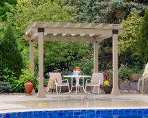 12x12 Vinyl Hollander Pergolas in almond vinyl on a patio by a pool with a table and two chairs.
